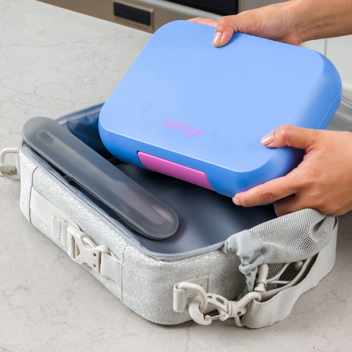 Person placing a blue and pink container into a gray suitcase on a light surface.