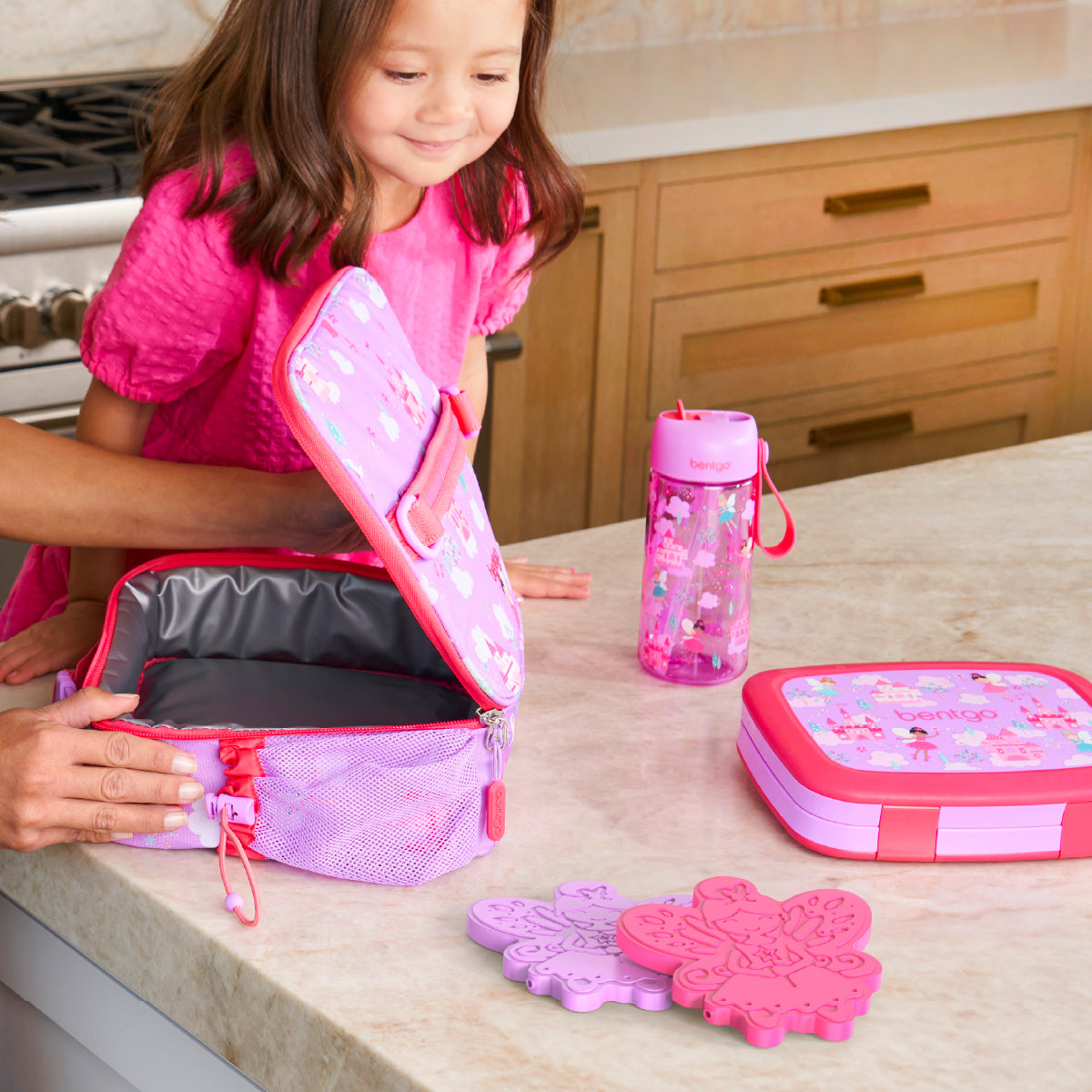 Child opening a pink lunch bag with matching water bottle and container on a kitchen counter.