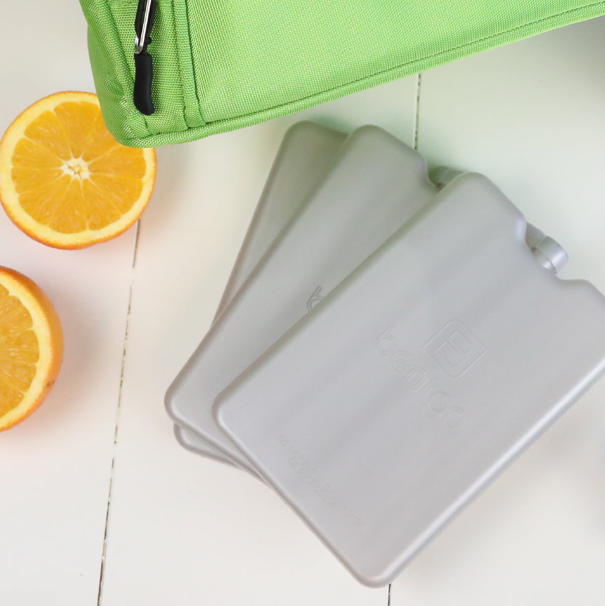 Three small white containers stacked on a light wooden surface with a green bag and orange slices in the background.