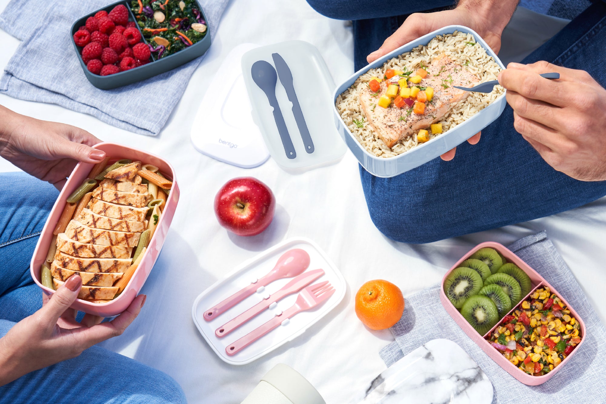 Hands holding various bento boxes with different foods on a light surface.