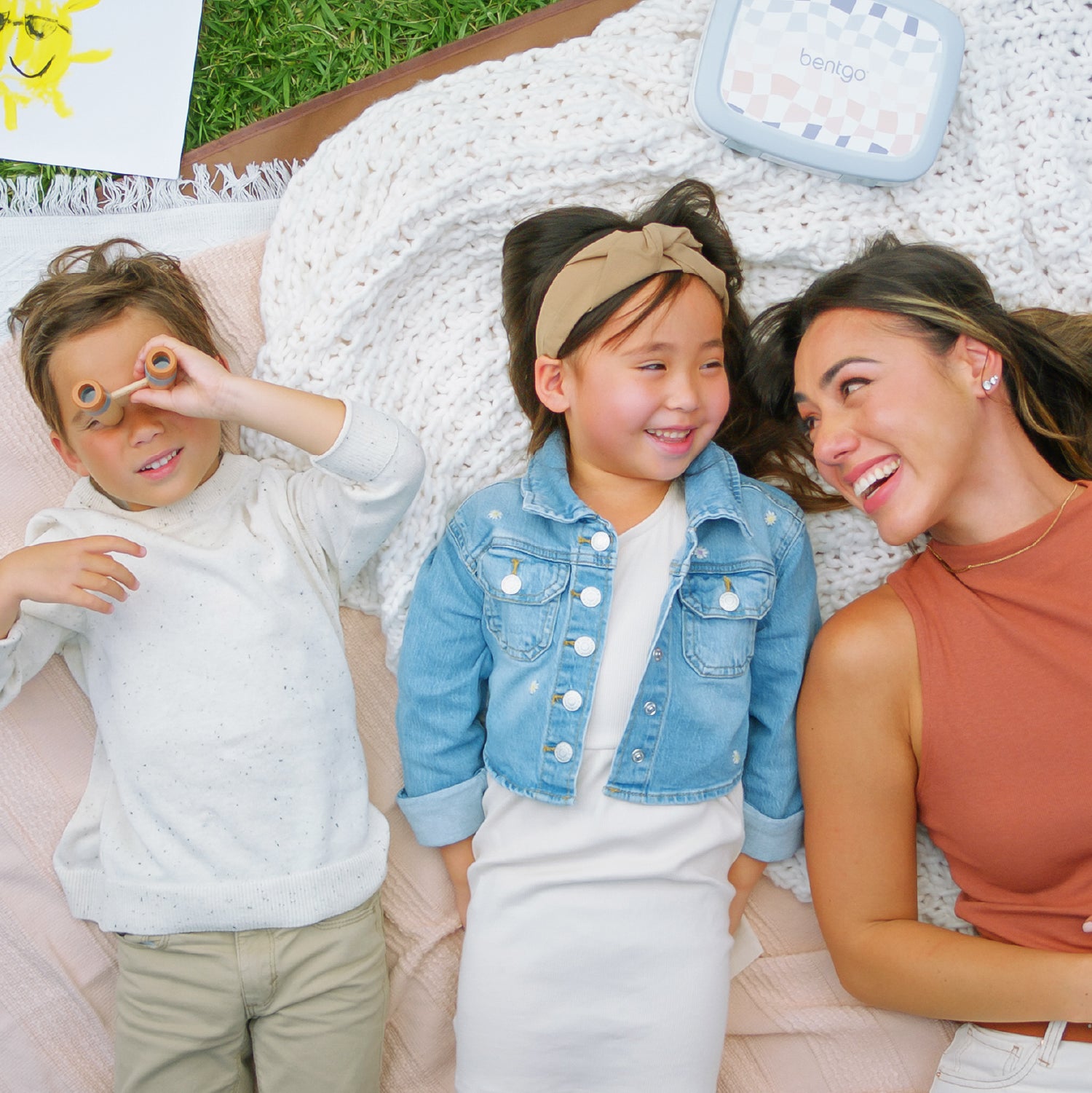 Woman and two children sitting on a blanket outdoors, with a baby bouncer in the background.