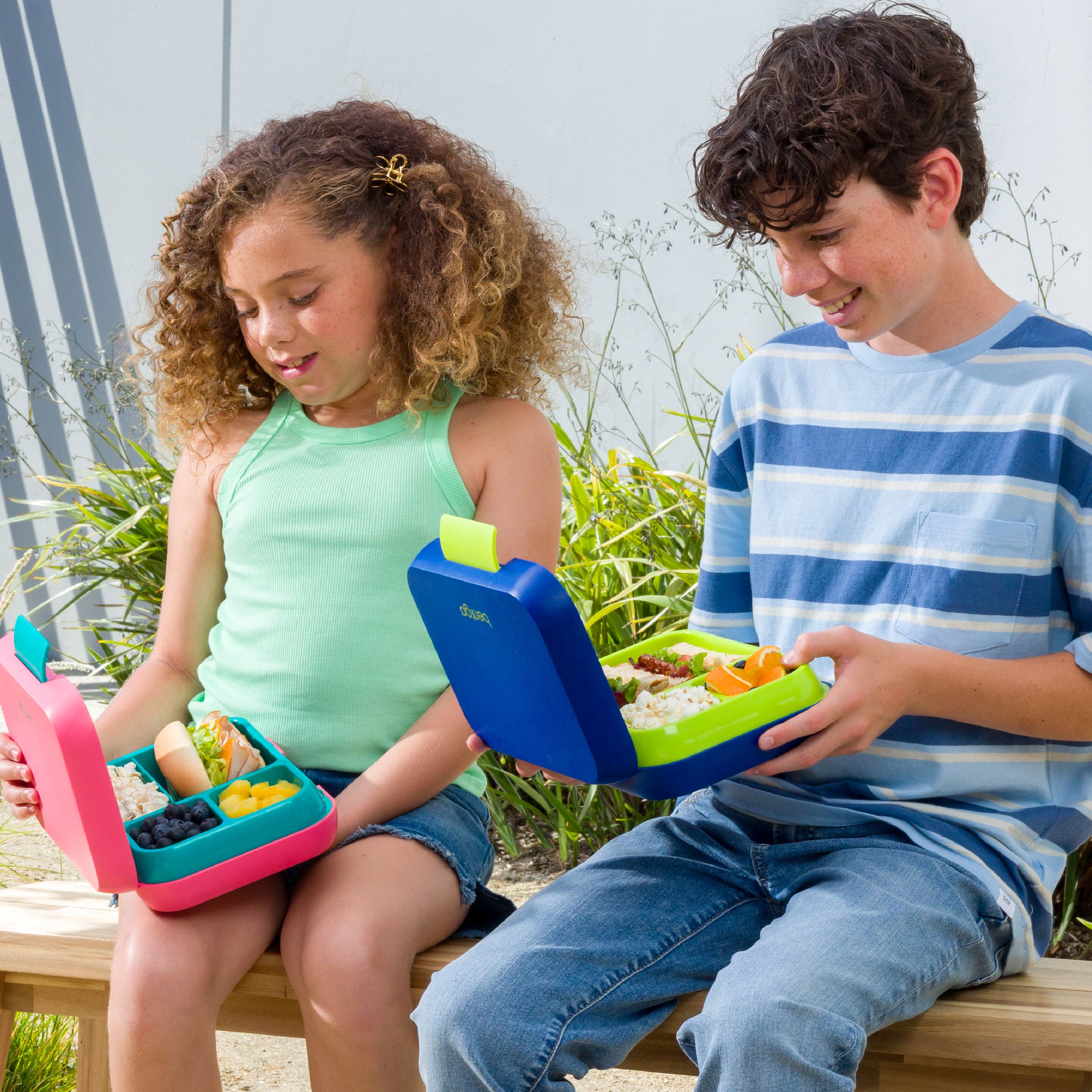 Two children sitting outdoors with lunch boxes containing snacks.