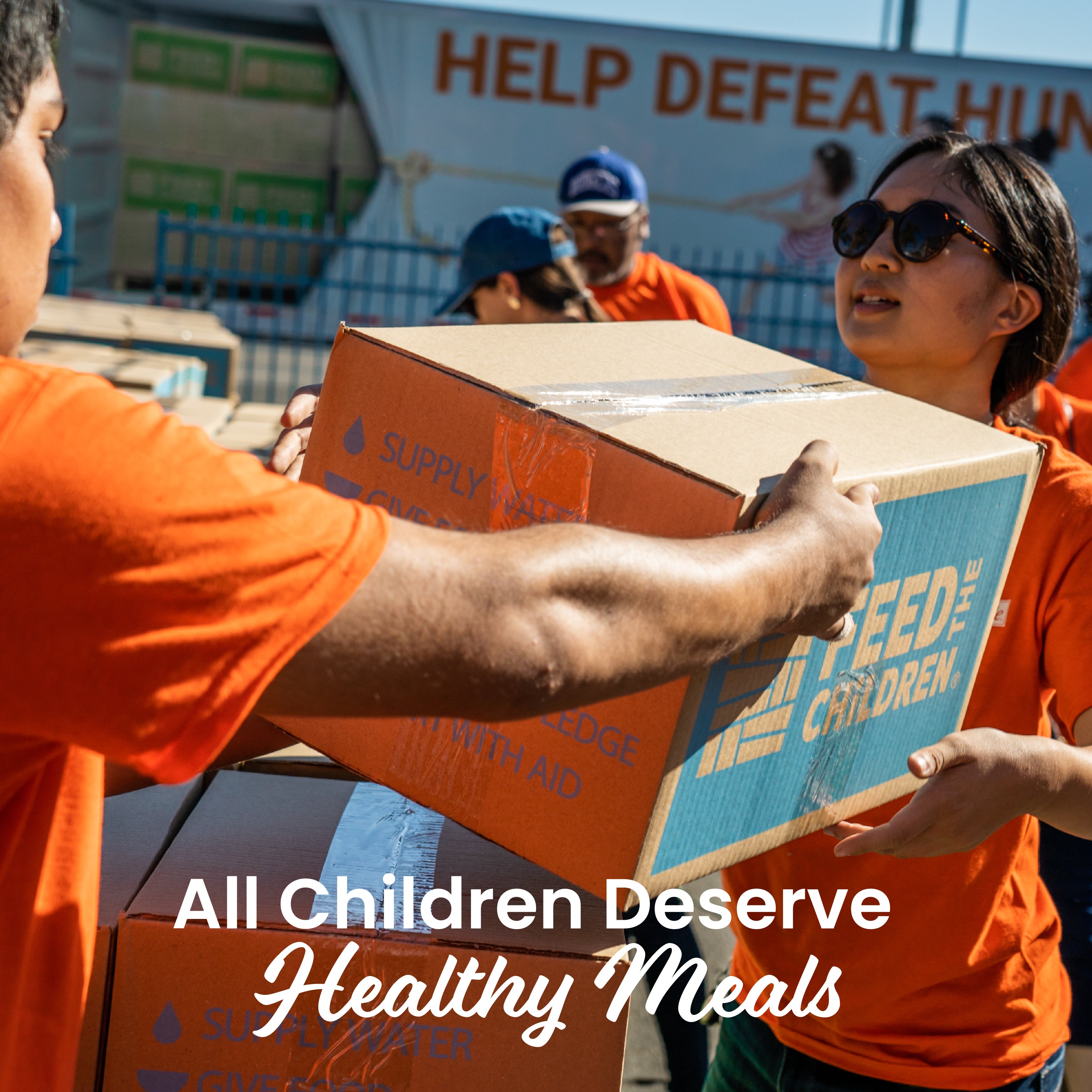 Two people in orange shirts handing over a box labeled 'Feed Children' with text 'All Children Deserve Healthy Meals'.